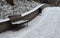 Snowy benches near the supporting concrete gray wall in the park. Paving and metal low fences protect ornamental flower beds from