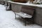 Snowy benches near the supporting concrete gray wall in the park. Paving and metal low fences protect ornamental flower beds from