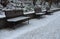 Snowy benches near the supporting concrete gray wall in the park. Paving and metal low fences protect ornamental flower beds from