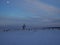 Snowy field with bales of hay and a lone tree at dusk with moon in the sky