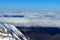 Snowy mountain tops peek out from under a layer of fluffy clouds. There is a snowy slope in the foreground. Beneath the