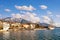 Snowy mountains and green seaside town under blue sky with white clouds . Montenegro, winter. View of embankment of Tivat city