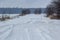 Snowy road in a field leading to pine forest. Winter road to nowhere in sunny day, snow-covered fresh car track. Car