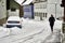 Snowy uncleared side street with high snow cover, next to which a car is parked and a dark-clad pedestrian is walking