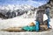 Snowy winter. A wooden board with a grey backpack, a blue thermos, a hat and a turquoise rope with blurred mountain background.