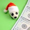 A soccer ball in a red Santa Claus hat next to the hundred dollar bills on a green background. Square frame for instagram. The