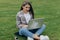Sociable young woman sitting on green grass in park with legs crossed during summer day while using laptop for video