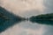 Soft focus. Silhouettes of fir hillside along mountain lake in dense fog. Reflection of coniferous trees in blue water. Alpine