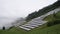 Solar park, photovoltaic power station located on a mountain slope in the Alps. Fog, rain, green pasture grass and mountains