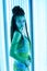 Solarium. Pretty girl with african braids in a dress for oriental dances sunbathing in a vertical sunbed. Blue neon glow