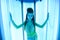 Solarium. Pretty girl with african braids in a dress for oriental dances sunbathing in a vertical sunbed. Blue neon glow