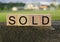 Sold text on wooden block on top of big stone with house background