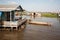 Solid floating house with a huge porch and children that sit in the shadow of the roof and the boat tied to the threshold