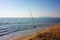 A solitary fishing rod rises from the ground on which it is planted on the sand of the beach and stands against the blue ocean sea