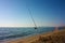 A solitary fishing rod rises from the ground on which it is planted on the sand of the beach and stands against the blue ocean sea