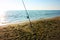 A solitary fishing rod rises from the ground on which it is planted on the sand of the beach and stands against the blue ocean sea