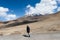 Solo Indian traveler in winter clothing standing alone in a remote mountain road. Snow capped mountain with floating clouds blue