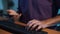 Some man in t-shirt is sitting at desk in dark room and actively typing text.