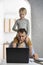 Son sits on his dads shoulders while he works at the laptop. Young father tries to work at home with the children