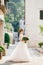 A sophisticated bride stands with a wedding bouquet in her hands near a white brick house in the old town of Perast