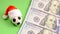 Souvenir soccer ball in a red Santa Claus hat next to three hundred US dollars on a green background. Christmas sporting events or