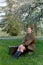 Soviet female soldier in uniform of World War II with suitcase sits on a stump near flowering tree