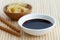 Soya sauce in white bowl on bamboo mat with chopsticks and pickled ginger in wooden bowl.