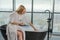 Spa relaxation. Young woman in spa bathrobe sitting at bath with foam in bathtub. Girl relaxing in bathroom at home