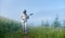 Spaceman in space suit with a guitar is walking on a beautiful green mountain meadow in foggy morning