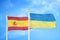 Spain and Ukraine two flags on flagpoles and blue cloudy sky