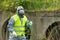 A specialist in a protective suit and mask holds in his hands a flask with a green reagent for examination at the site of the