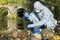 A specialist in a protective suit and respirator analyzes the water in the river near the existing plant, using a flask and a