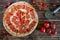 Spicy pepperoni and salami pizza lies on a wooden surface of pine planks next to a special circular knife, tomatoes and chili.