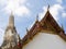 The spire of the Wat Arun Temple in Bangkok, Thailand, with intricate roof details in red an gold and a blue sky with clouds