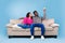 Split system. Young black couple with remote relaxing under air conditioner, sitting on couch over blue background