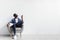 Split system. Young black man with remote turning on air conditioner, sitting in armchair against white wall, mockup