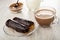 Spoon in bowl of cocoa powder, pitcher with milk, eclairs with chocolate in plate, cup of cocoa with milk on table