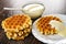 Spoon in bowl with condensed milk, stack of waffles, plate with biscuit waffle poured condensed milk