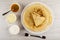 Spoon in bowl with honey, bowls with sour cream, condensed milk, pancake on stack of pancakes in dish, fork on table. Top view