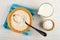 Spoon in bowl with raw rice on napkin, jug of milk, salt cellar on wooden table. Top view