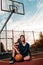 Sports and basketball. A young teenager in a black tracksuit poses with a basketball on a sports field while sitting on the ground