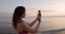 Sporty attractive woman standing with smartphone and shooting sea with sunset or sunrise on beach