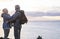 Sporty elderly couple on hike on ocean cliffs. On the horizon the profile of an island and a ship. Cloudy sky at sunset