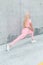 Sporty happy young woman athlete model in stylish pink tracksuit with white shoes doing stretching near gray concrete wall