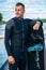 Sporty man in wetsuit standing on pier leaning on wakeboard and looking away
