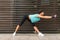 Sporty young woman doing wide-legged forward bend exercise holding her arms straight on the city street.