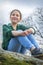 In the spring afternoon, a girl with a scythe sits on the edge of a cliff in blue jeans and a green shirt, side view