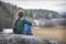 In the spring afternoon, a girl with a scythe sits on the edge of a cliff in blue jeans and a green shirt, side view