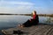 In the spring, a beautiful woman in a burgundy embroidered dress rests on a wooden pier near the calm water