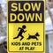 Square Black and yellow Slow Down Kids And Pets At Play sign against gray wooden pole